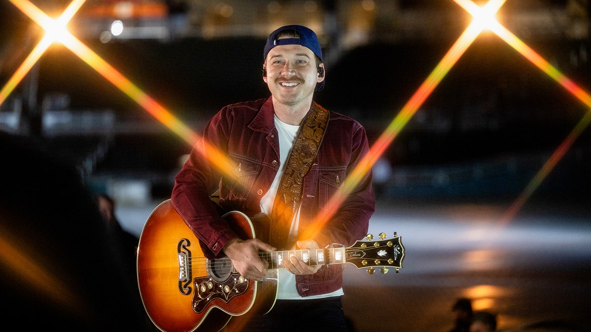 Morgan Wallen in a red jacket playing the guitar with a backwards hat