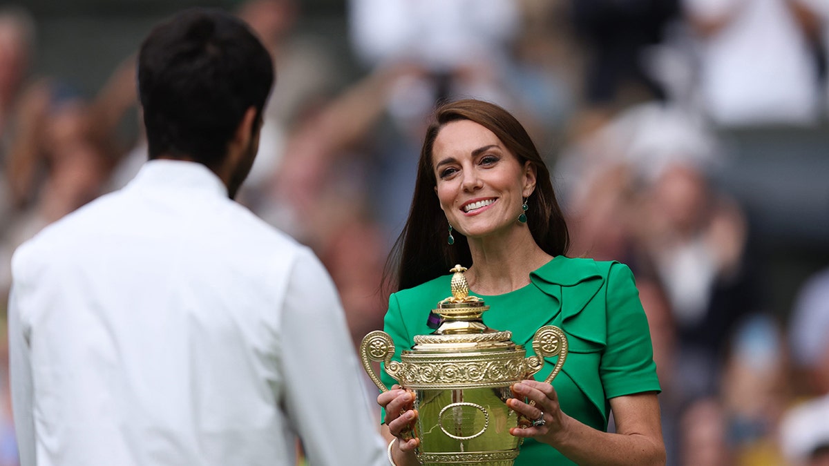 Kate Middleton in a bright green dress holds a silver trophy and presents it to Carlos Alcaraz