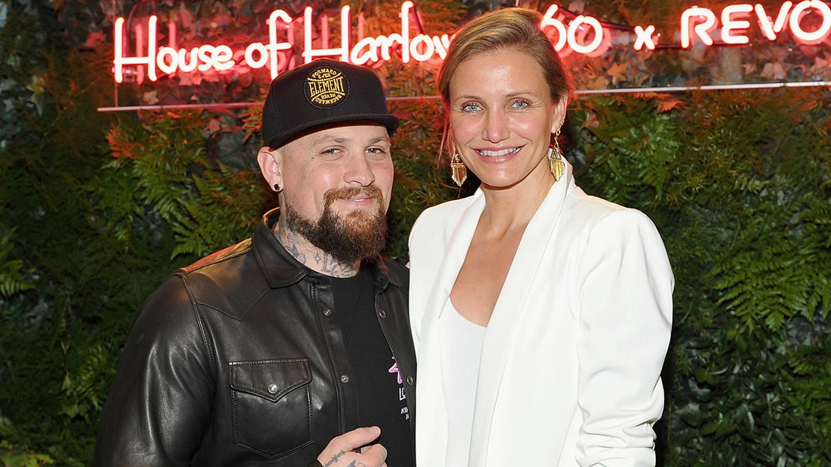 Benji Madden in a leather jacket and black hat smiles next to wife Cameron Diaz in a white blazer