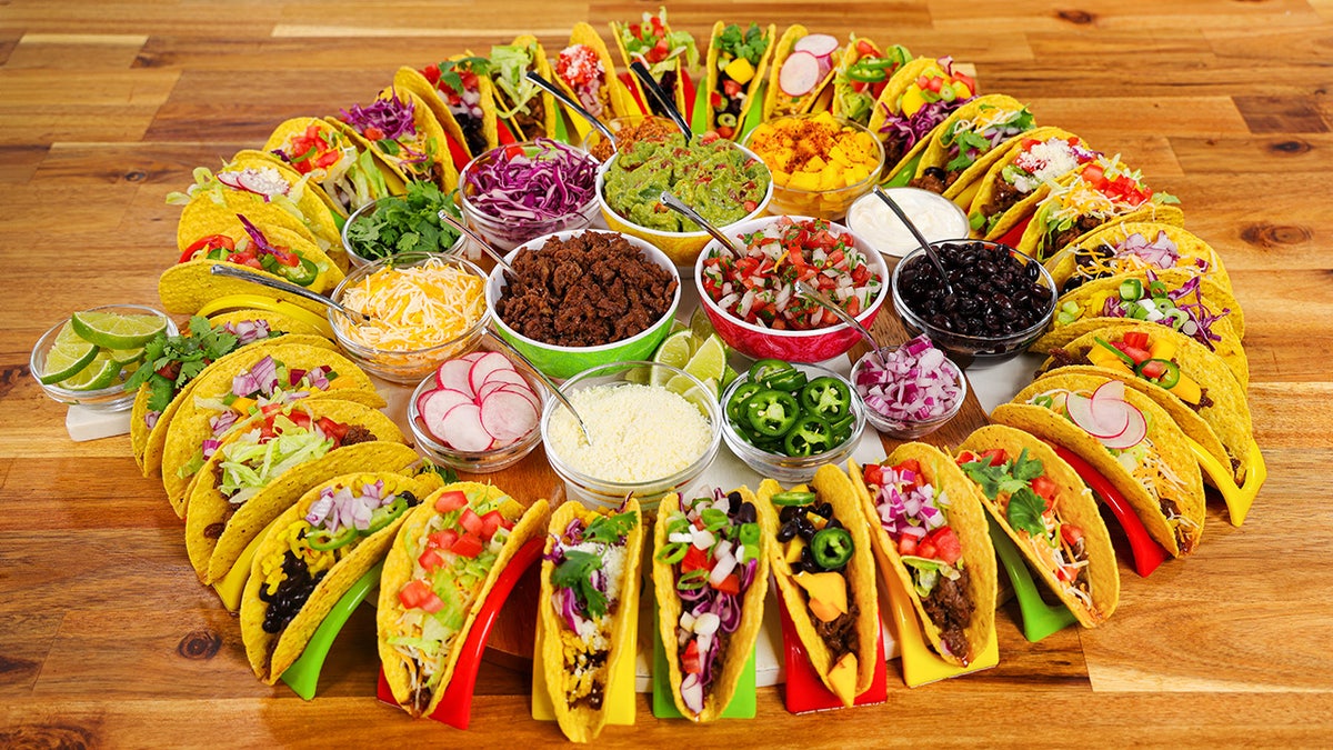 A taco board consisting of a circle of filled hard taco shells with various taco toppings in the middle.