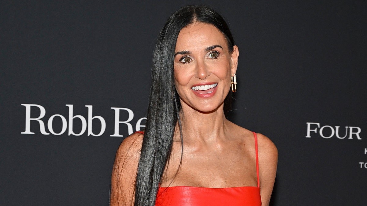 Demi Moore in a red latex strappy dress looks to her left on the carpet as she smiles