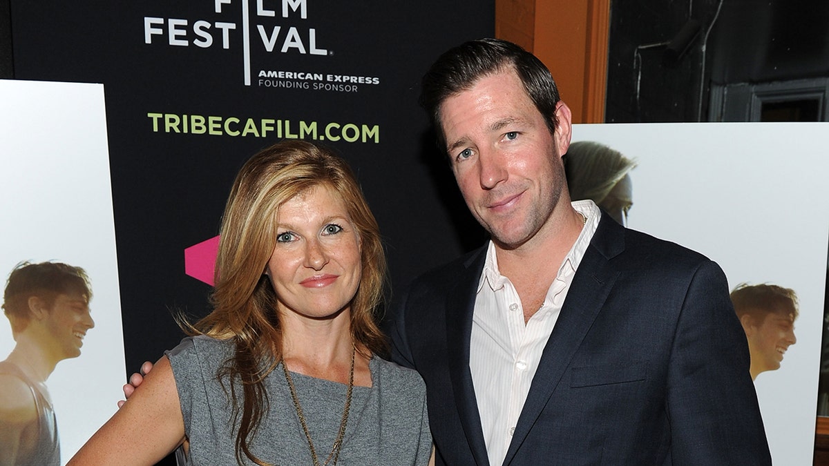 Ed Burns smiling in a navy blazer and a white shirt next to Connie Britton who is wearing a grey sweater.