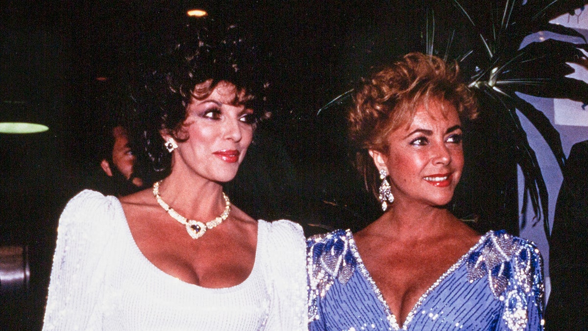 Joan Collins in a white gown standing next to Elizabeth Taylor, who is wearing a light blue sparkling gown.