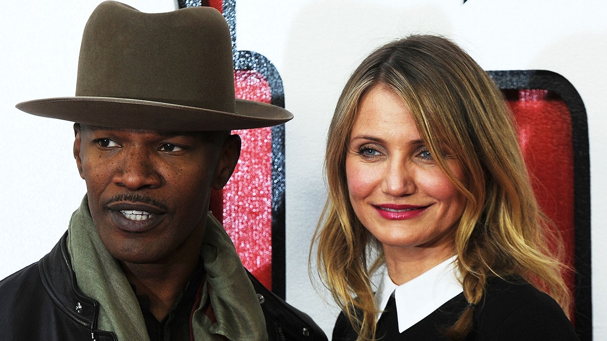 Jamie Foxx in a green scarf and hat smiles on the carpet with and Cameron Diaz in a black sweater and white collar shirt