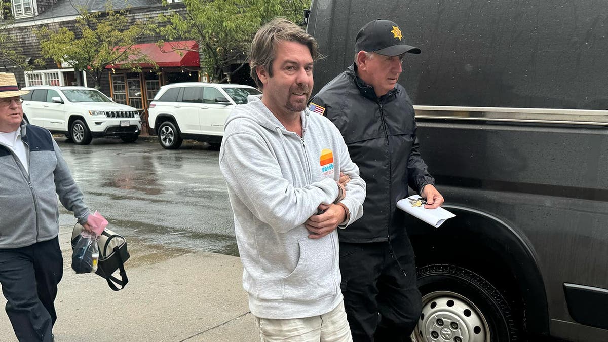 A former advisor to John Kerry is handcuffed and wearing a grey hoodie as he is escorted by law enforcement on a rainy day