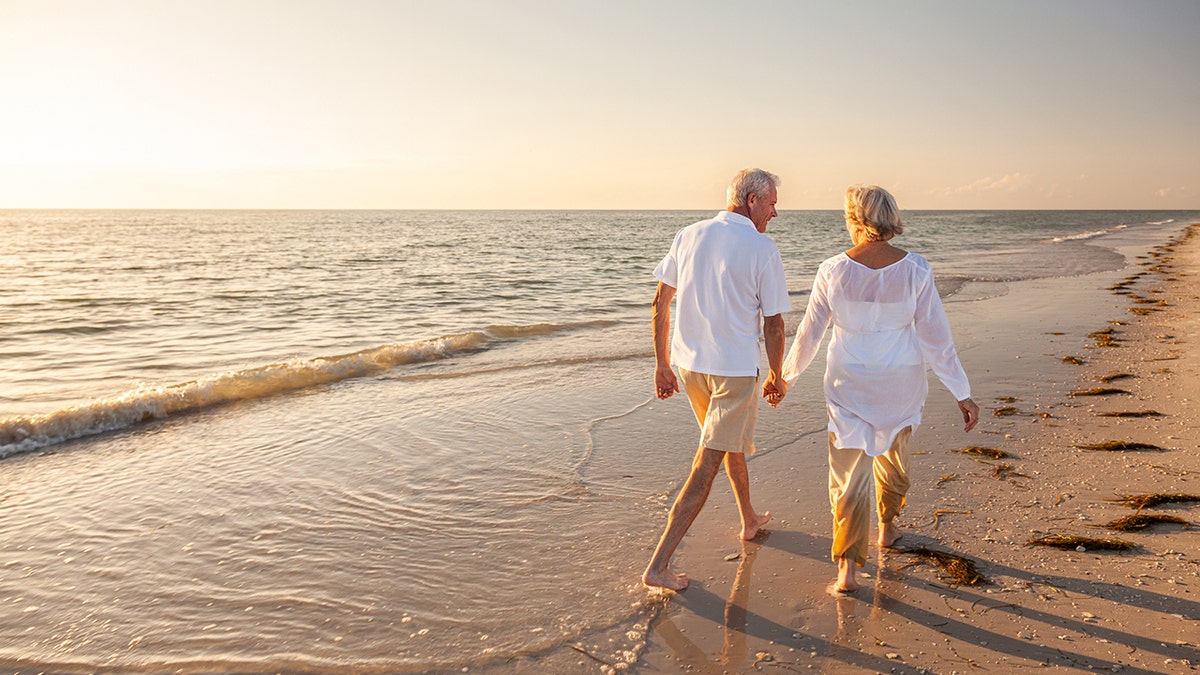 Happy senior man and woman walking and holding hands on a beach at sunset