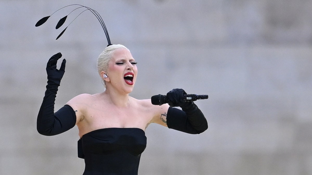 Lady Gaga in a strapless black gown with long black gloves sings into the microphone passionately with a feather sticking out of her hair