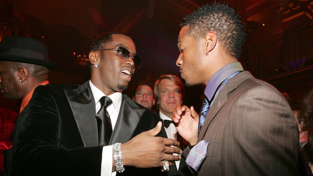 Sean 'Diddy' combs in a black suit and tie talks with Marlon Wayans in a brown suit at an event in NYC