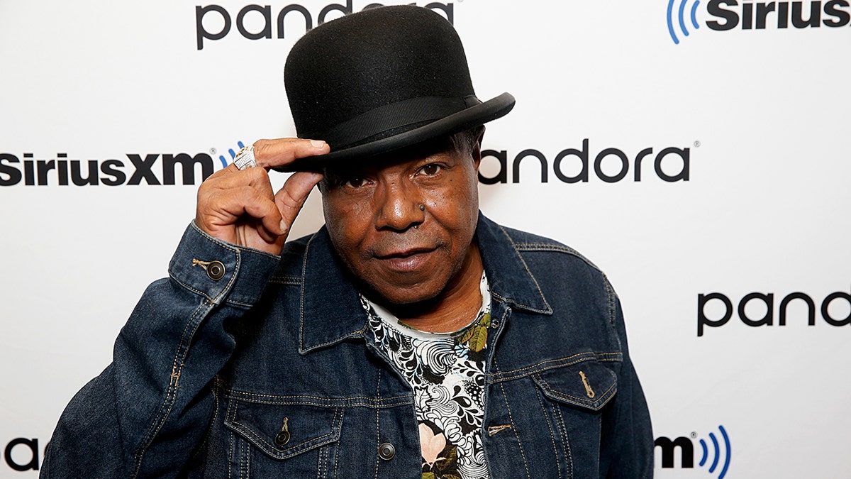 Tito Jackson in a. jean jacket looks at the camera as he pulls down his black hat