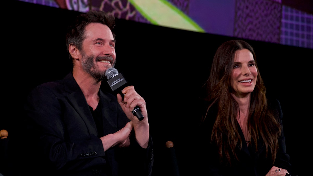 Keanu Reeves smiles on stage as he holds a microphone to his mouth sitting next to smiling Sandra Bullock, both in black