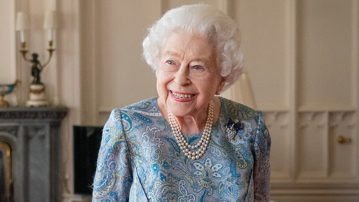 Queen Elizabeth smiles and looks off in the distance in a blue patterned dress and strings of pearls at Windsor Castle