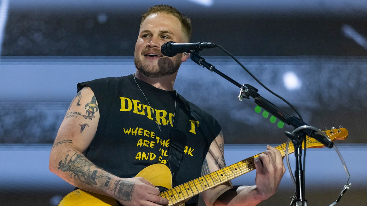 Zach Bryan in a black cut-off tank top with yellow writing and a yellow guitar on stage