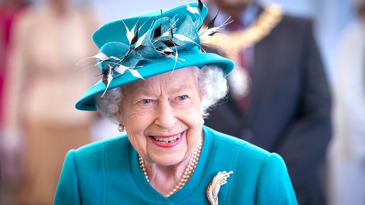 Queen Elizabeth II smiles in a teal jacket and matching hat with feathers sticking out