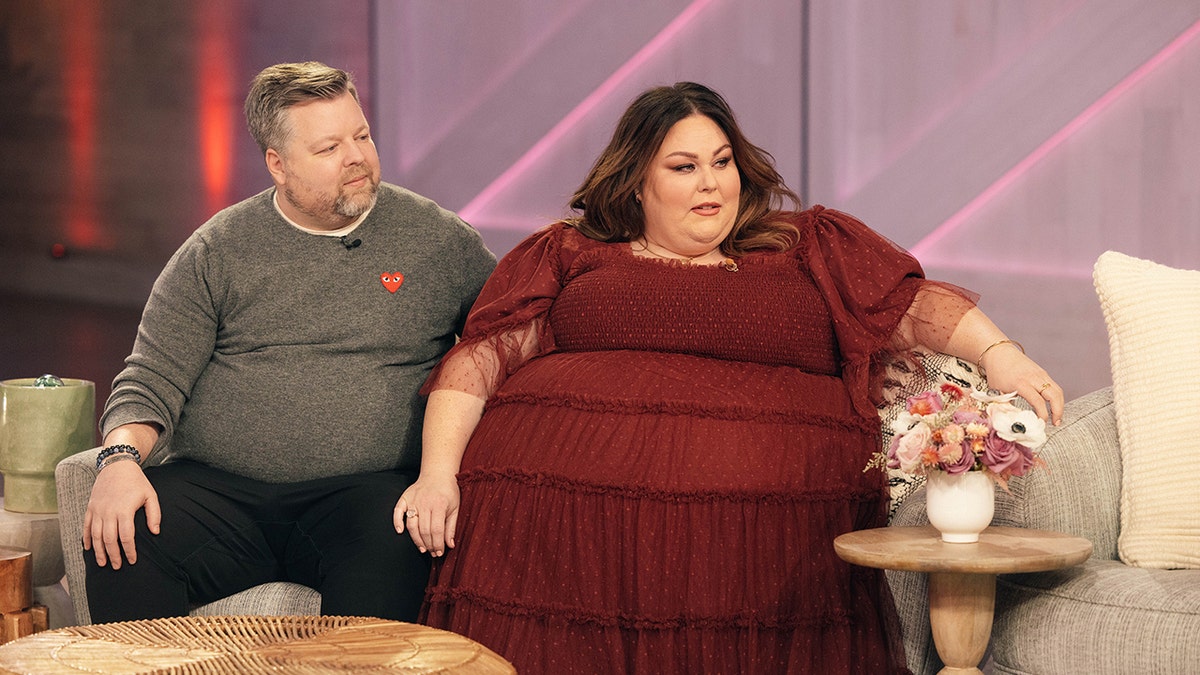 Chrissy Metz in a long maroon ruffled dress rests her hand on her boyfriend Bradley Collin's knee wearing a grey sweater while on 