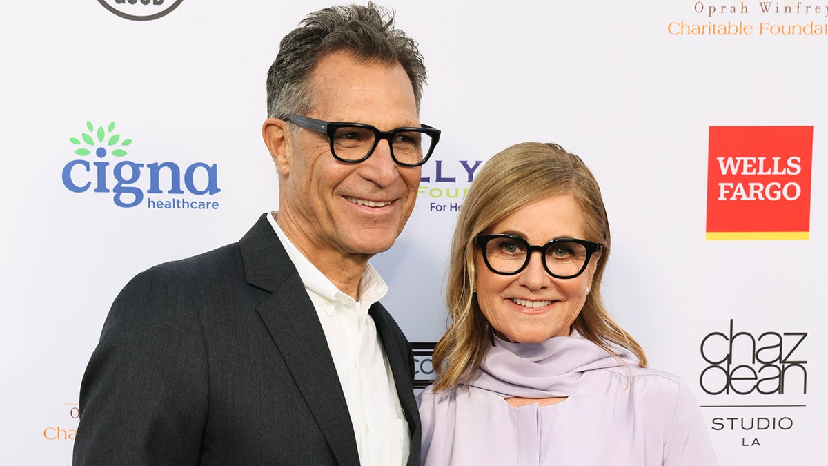 Maureen McCormick in a lavender blouse and black sunglasses standing next to her husband in a dark blazer, a white shirt and black sunglasses.