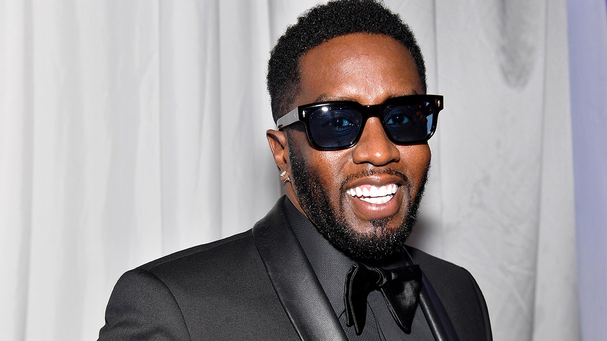 Sean Diddy Combs in a black tuxedo and black sunglasses smiles wide