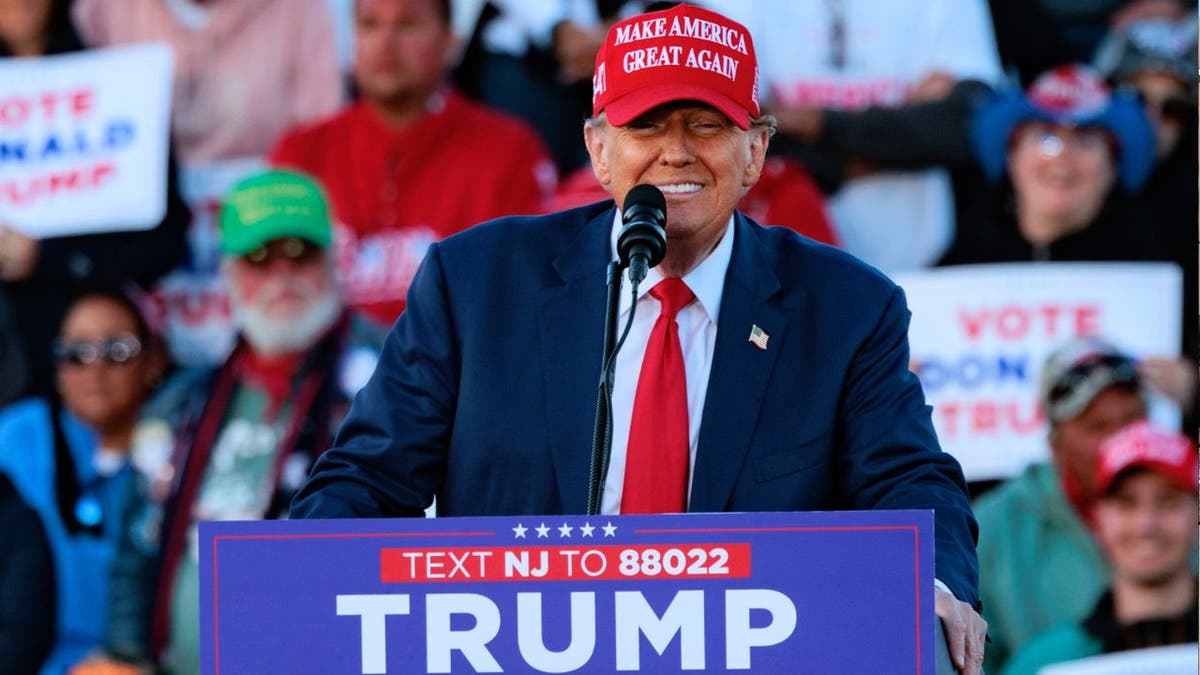 Trump in red MAGA hat smiling at lectern at rally