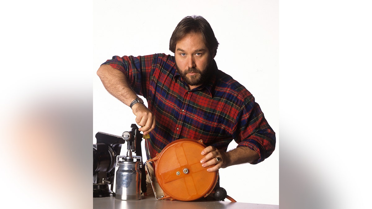 Richard Karn in a red plaid shirt holds some type of machinery in character for 