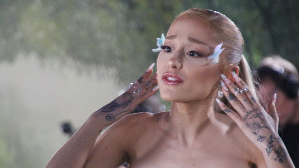 Ariana Grande puts her hands up to her ears and looks distressed with white wings glued to the side of her face