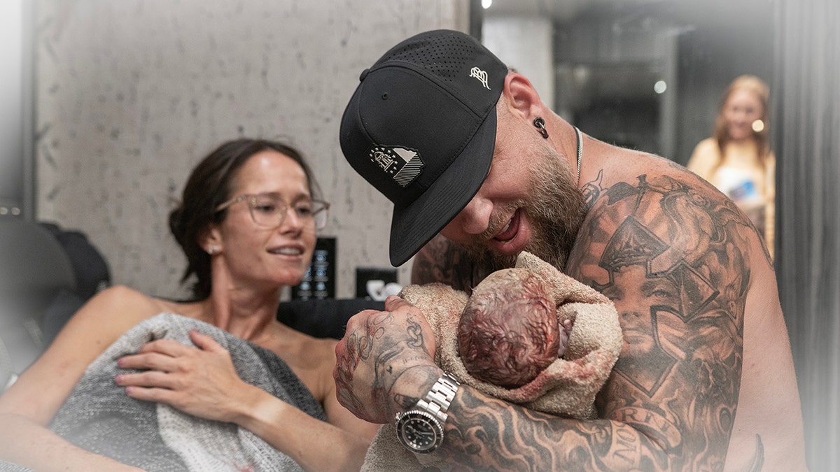 Brantley Gilbert holds his new baby wearing a black hat while his wife beneath a blanket looks on