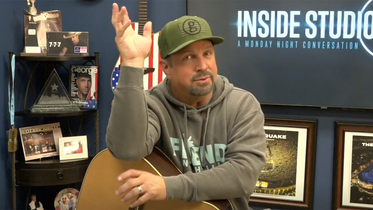 Garth Brooks in a sweatshirt and green baseball cap puts his hand in the air as he rest the other over his guitar