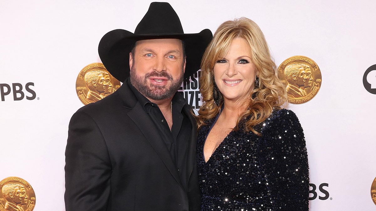 Garth Brooks in a black suit and black cowboy hat soft smiles with his wife Trisha Yearwood in a red cheetah dress