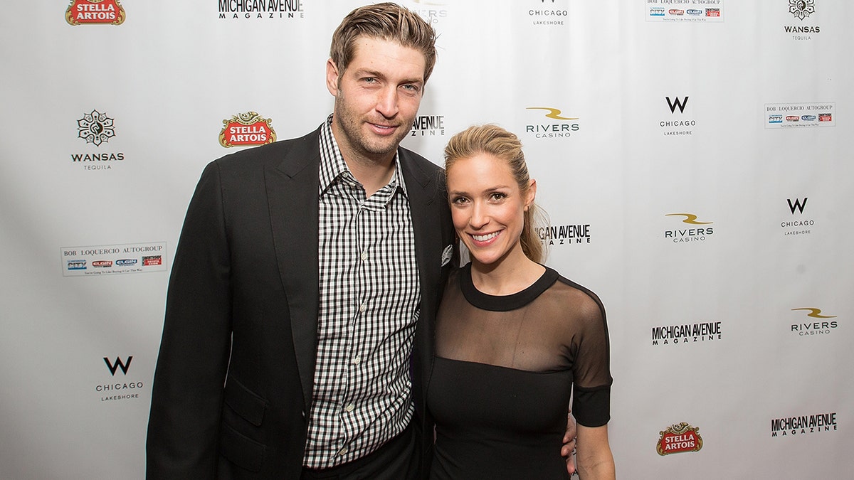 Jay Cutler ina black suit and checkered shirt smiles on the carpet with Kristin Cavallari in a black dress with a mesh top