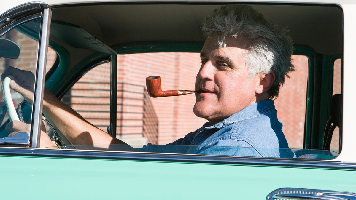 Close up of Jay Leno sitting in a car smoking a pipe