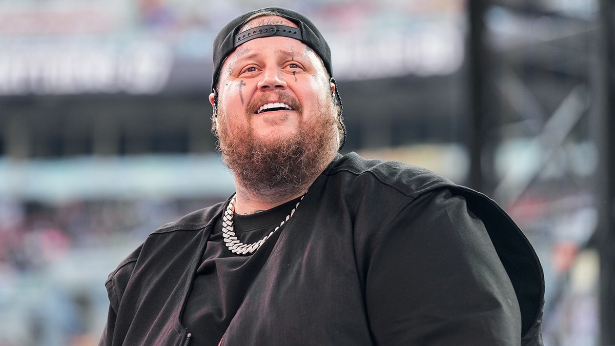 Jelly Roll in a black shirt and black backwards hat looks up and smiles at crowd