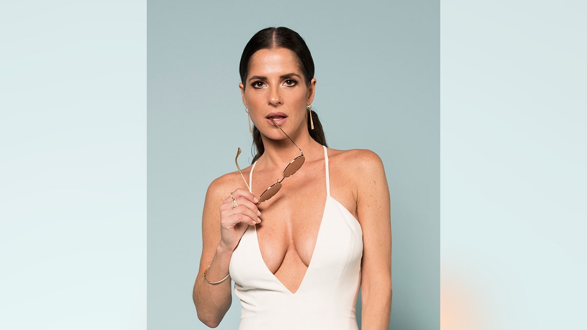 Kelly Monaco in a plunging white outfit holds her sunglasses seductively