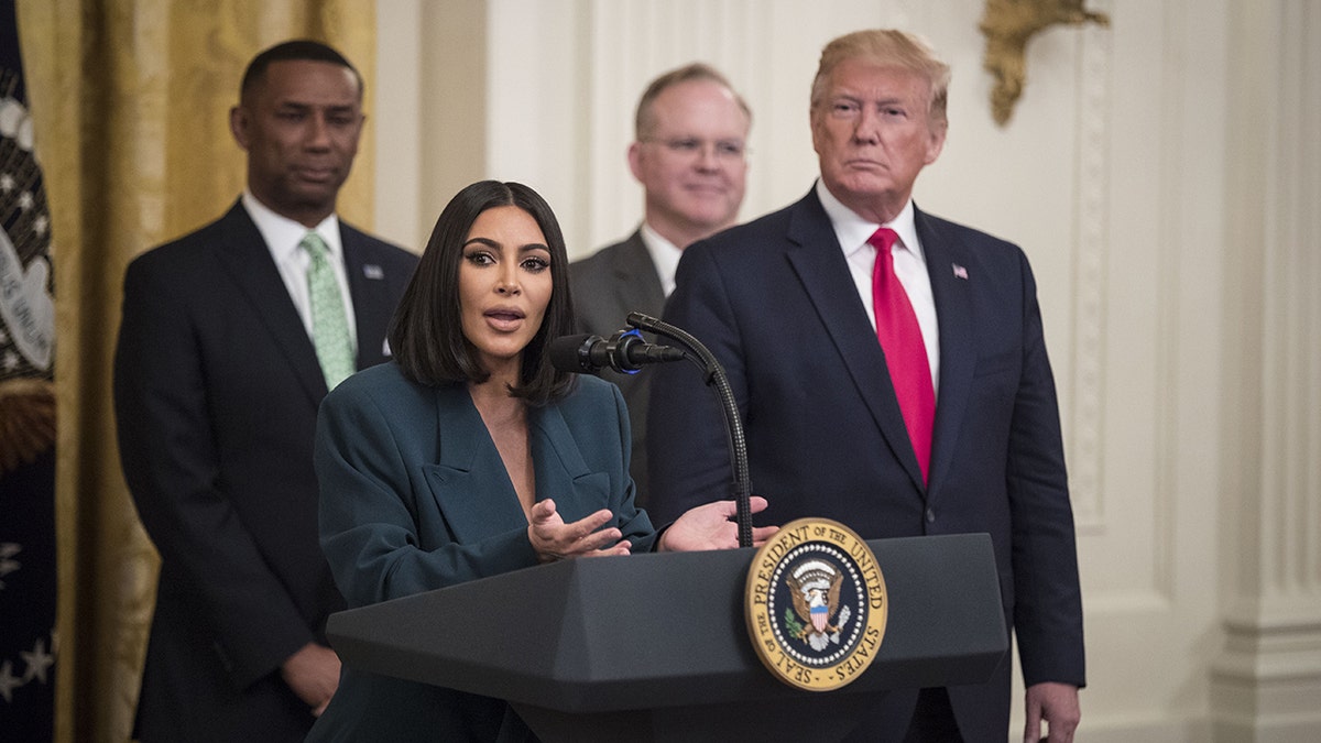 Kim Kardashian in a dark teal suit stands behind the podium at the White House with former President Trump in a dark suit and red tie standing to her left