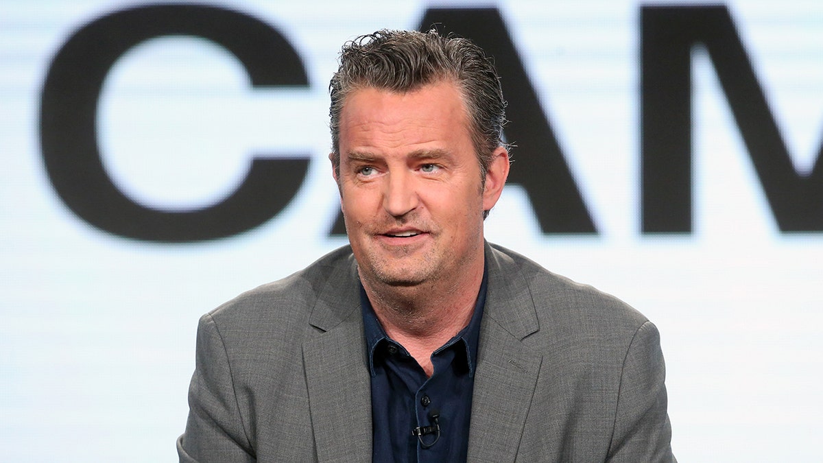 Matthew Perry in a grey suit and dark shirt looks off in the distance on stage