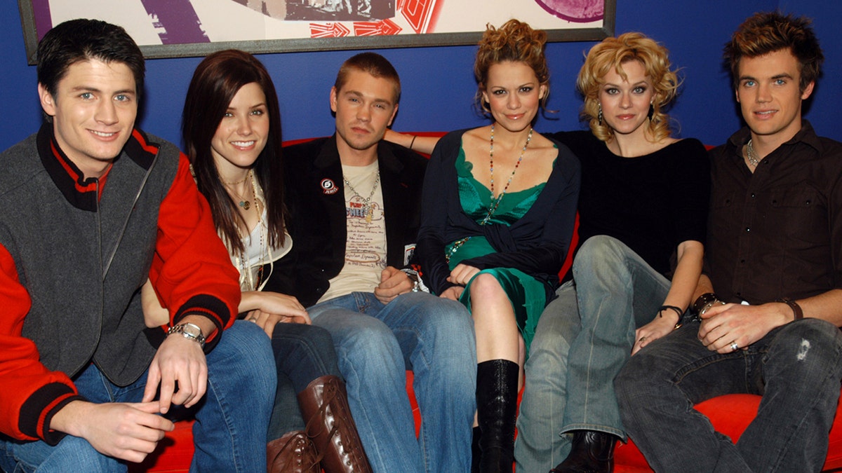 James Lafferty, Sophia Bush, Chad Michael Murray, Bethany Joy Lenz, Hilarie Burton and Tyler Hilton all sit side by side on a red couch