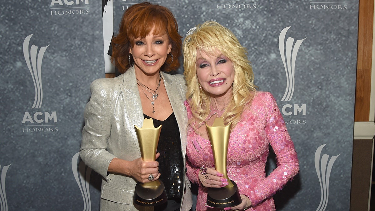 Reba McEntire in a gold sparkly suit and black shirt holds an ACM awards with Dolly Parton in a bubblegum pink dress, also holding an ACM