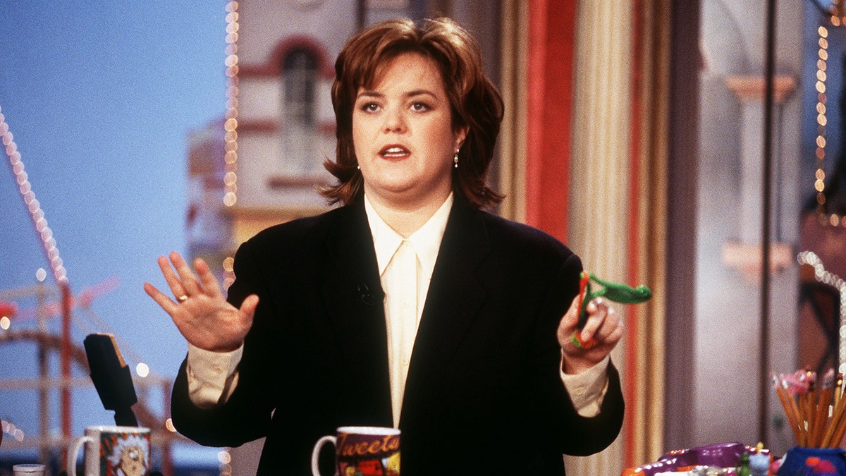 Rosie O'Donnell in a black suit puts her hands up as she hosts her talk show 