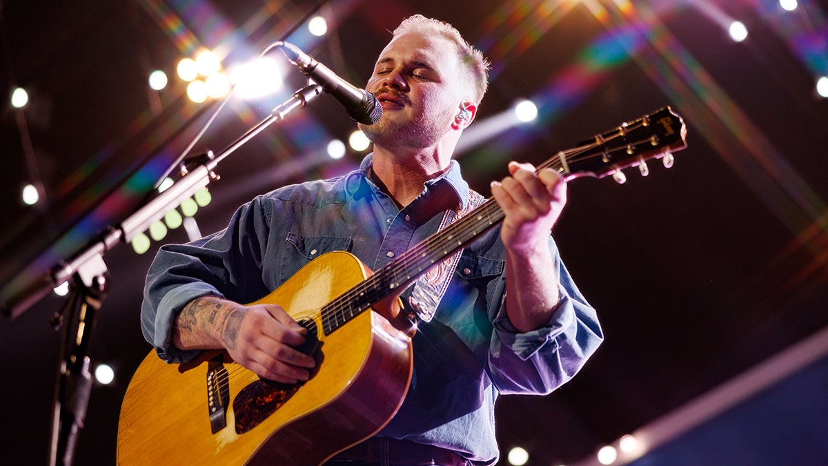 Zach Bryan in a blue denim shirt sings into the microphone while strumming the guitar