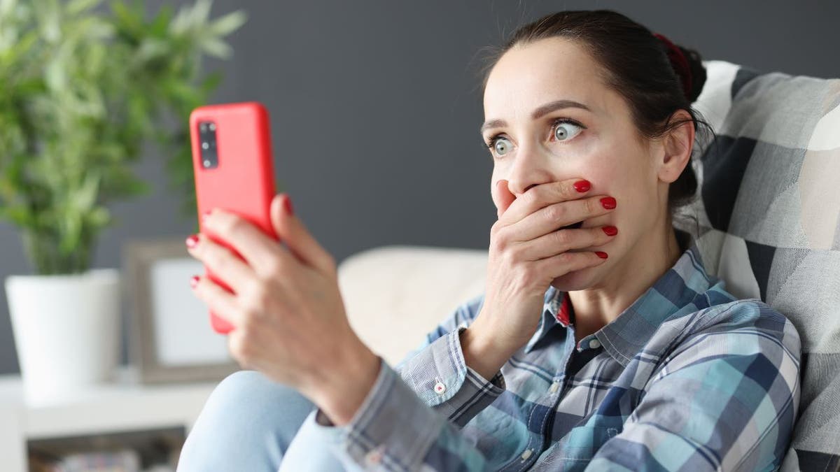 Woman covering mouth with hand, eyes wide in shock while holding up iPhone