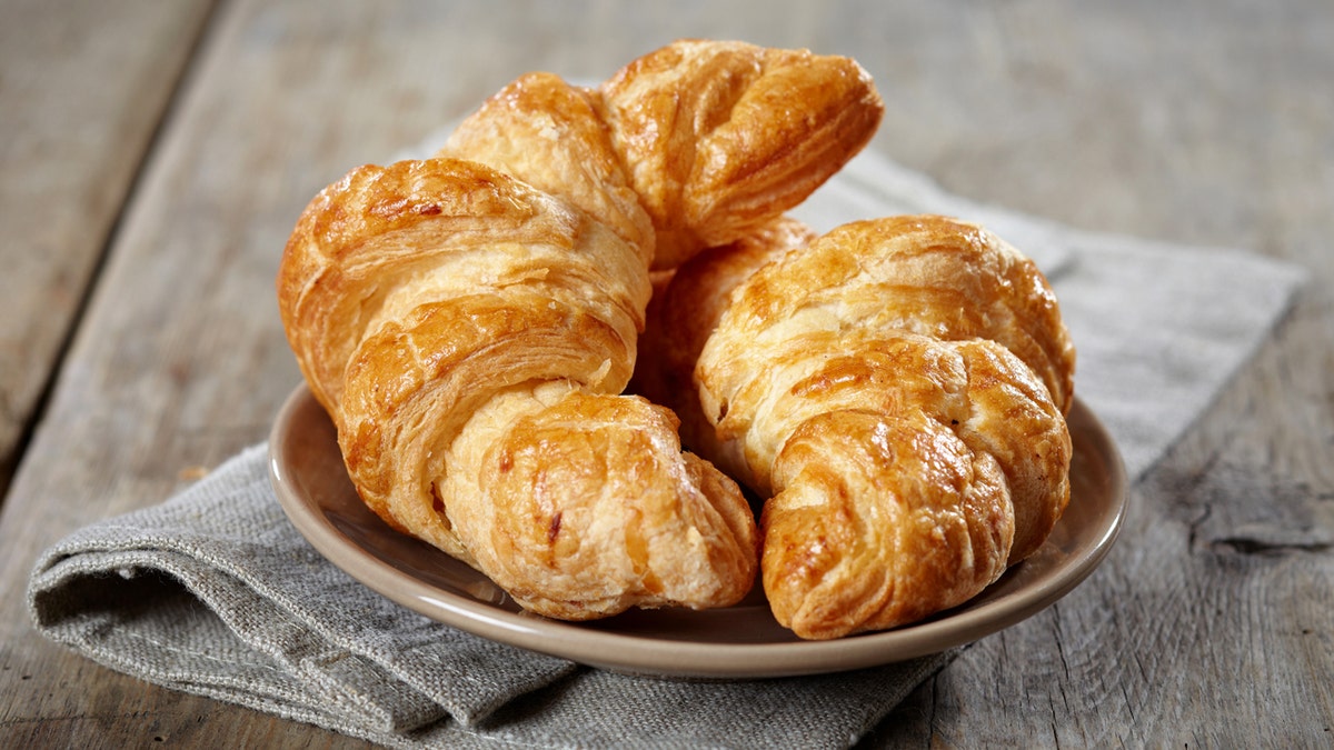 Two butter croissants rest on a plate beneath a cloth napkin on a wooden table.