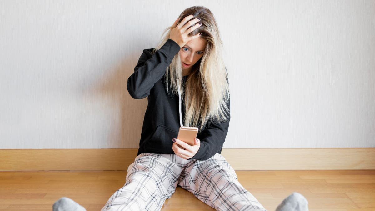 Woman places hand on head in shock with eyes wide and mouth open looking down at her phone