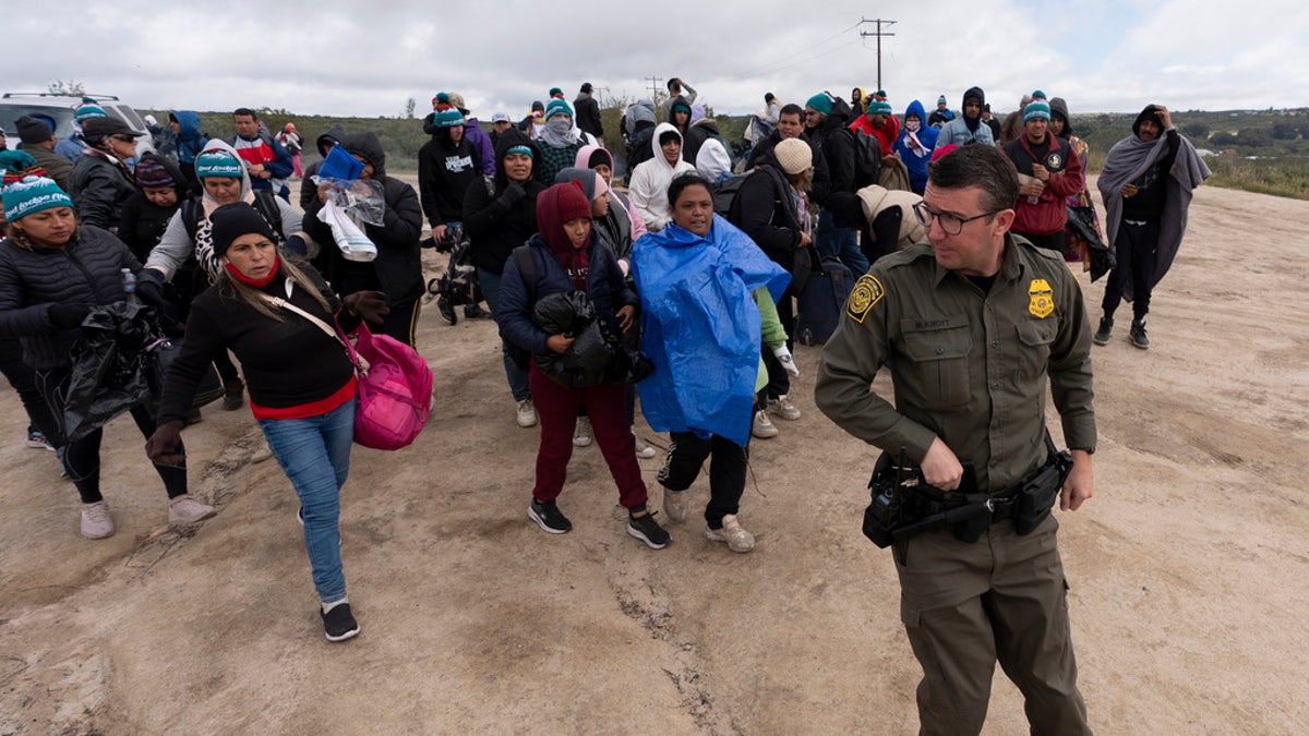 People seeking asylum, including a group from Peru, walk behind a Border Patrol agent towards a van 