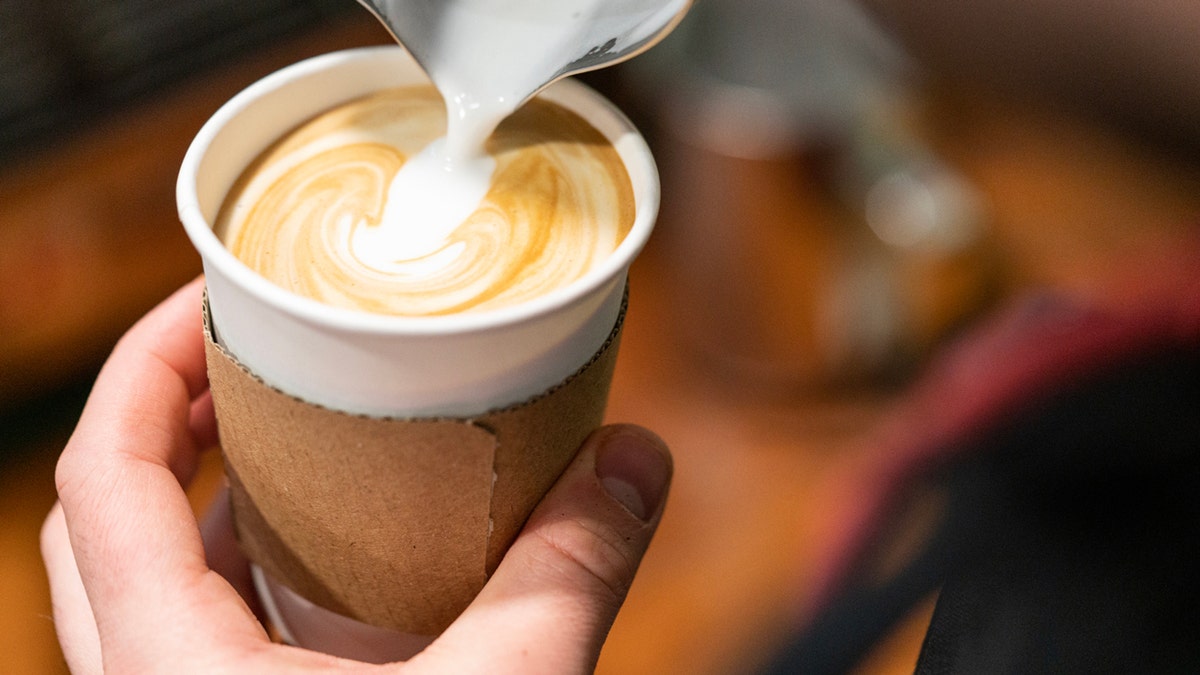 A barista holds a cup of latte as milk is being poured into it.