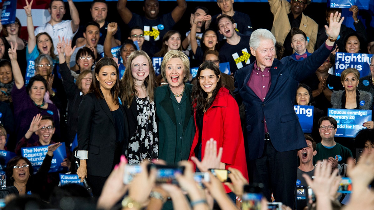 Eva Longoria in black poses with Chelsea Clinton in a black patterned dress, Hillary Clinton in a green suit, America Ferrera in a red jacket, and Bill Clinton looking out towards the audience in a blue suit and purple shirt