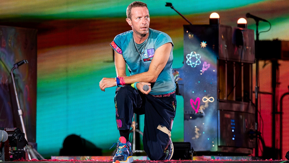 Chris Martin kneels on stage in a turquoise shirt and looks to his left