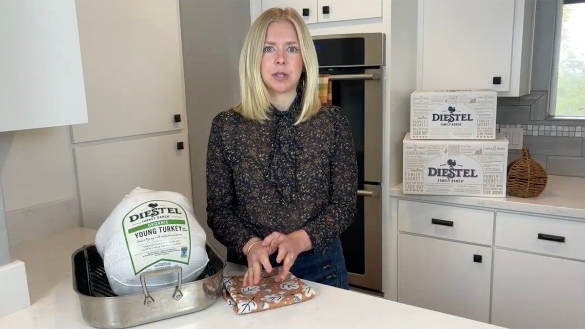 Blonde woman standing next to packaged turkey in kitchen.