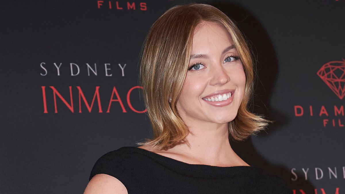 Sydney Sweeney smiles at the camera in a short-sleeve black dress