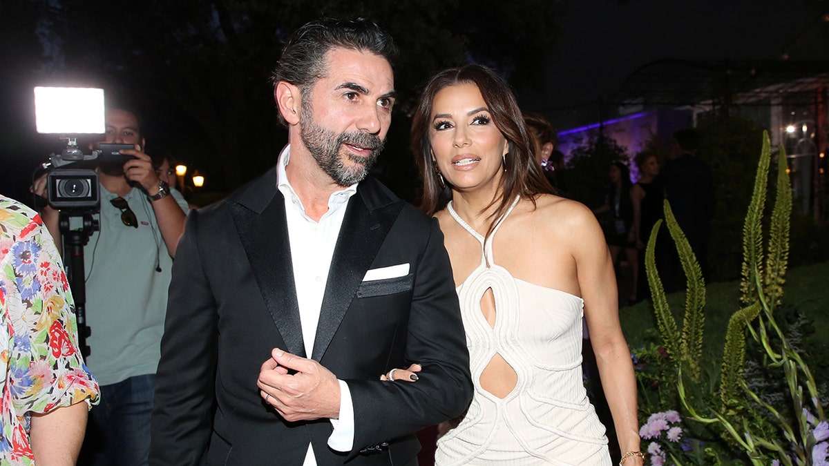 José Bastón in a black jacket is held onto by wife Eva Longoria in a white halter dress with cutouts as they walk in Spain