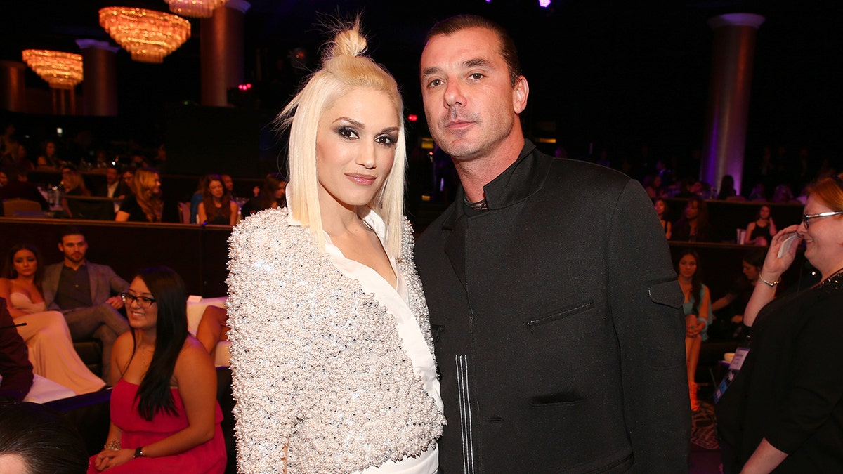 Gwen Stefani with a tiny knot bun on her head and pearl-like sweater looks at the camera with husband Gavin Rossdale in a black coat