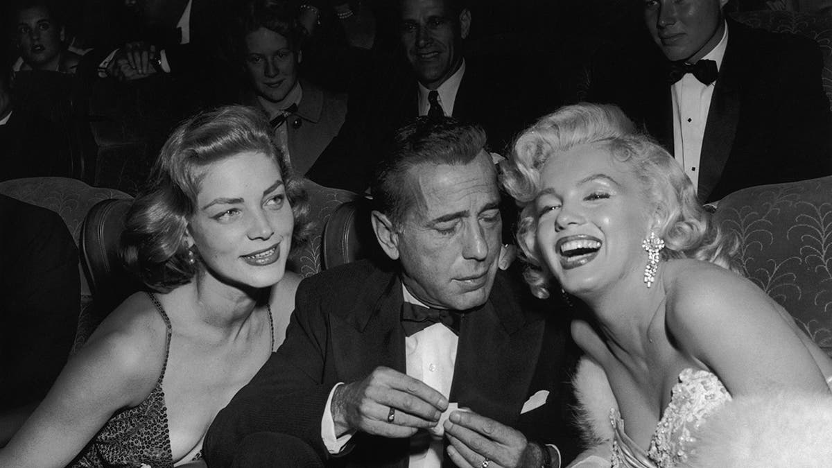 A close-up of Humphrey Bogart in a tux sitting in between Lauren Bacall and Marilyn Monroe.