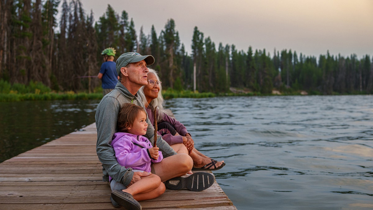 couple sit on a dock with their granddaughter while watching the sunset over a lake