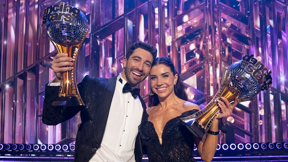 Joey Graziadei in a tuxedo and Jenna Johnson in an off the shoulder black gown both hold Mirrorball trophies in the air